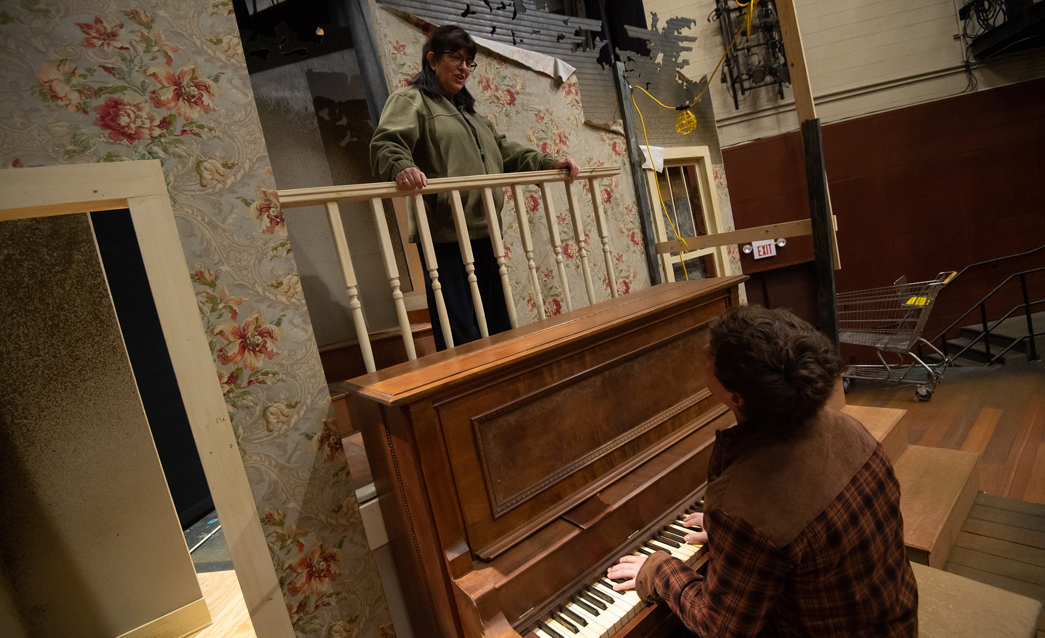 Maxwell Glasser plays the piano while looking up at Rene Pulliam, who is standing on a staircase landing above the piano. 
