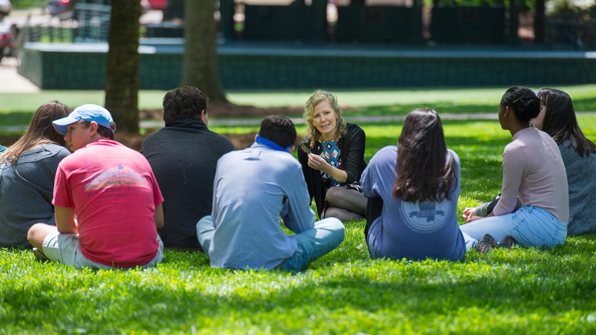 Professor Katie McKee and a group of 7 honors English students sit on the grass in the Grove, having a discussion together